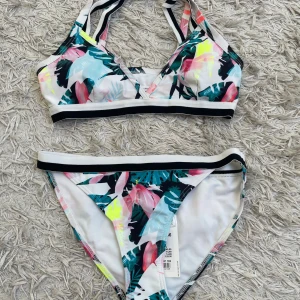 Bikini från SOC - Superfin bikini från SOC i storlek 36 med tropiskt blommigt mönster i vitt, rosa, blått, grönt och inslag av neon. Både överdel och underdel har breda svarta och vita resårband och sportiga axelband. Helt oanvänd med prislappen på.