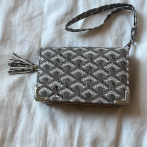 Gråvit mönstrad plånbok  - Snygg plånbok i grått och vitt med geometriskt mönster, silverfärgade metalldetaljer och en dekorativ tofs. Plånboken har flera fack för kort, myntficka med dragkedja och transparenta fönster för ID. Praktisk handledsrem för enkel användning.