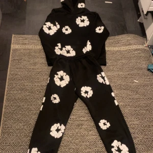  Svart   Denim Tear tracksuit - Svart  Denim Tears traksuit  med stora vita bummul  över hela tröjan och byxorna . Tröjan har huva och en relaxed passform, perfekt för en cool och avslappnad stil. Materialet känns mjukt och skönt mot huden. Samma material för byxorna.