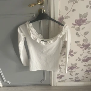 Vit långärmad topp med volang - Säljer en vit långärmad topp med bred volang runt den vida, runda urringningen. Toppen har en feminin och stilren look, perfekt att styla med jeans eller kjol. Materialet känns mjukt och stretchigt.