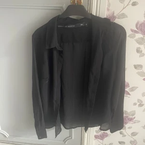 Svart transparent skjorta S - Svart, tunn och transparent skjorta från The Black Label Collection. Skjortan har klassisk krage, långa ärmar med manschett och en loose passform. Perfekt för lager-på-lager och en stilren look.