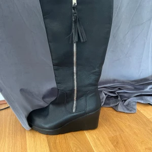 Svarta stövlar med kilklack och dragkedja - Säljer ett par svarta stövlar med snygg kilklack och grov sula. Skorna har dragkedja på sidan och dekorativ tofs. Tillverkade i syntetmaterial med en stilren look som passar till många outfits.