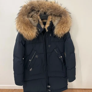 Svart dunjacka med päls från Cedrico - Säljer en svart dunjacka från Cedrico med stor fluffig päls runt huvan. Jackan har dragkedja, knappar och flera fickor med snygga detaljer. Perfekt för kalla vinterdagar och riktigt varm. Klassisk lång modell med justerbar midja.