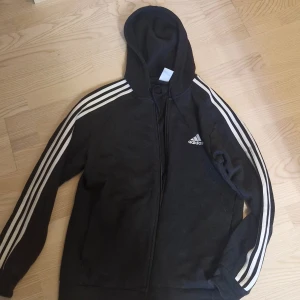 Svart Adidas hoodie med vita ränder - Svart hoodie från Adidas med klassiska vita ränder längs ärmarna och Adidas-logga på bröstet. Tröjan har huva, dragkedja framtill och långa ärmar. Perfekt för en sportig och avslappnad stil.. Katt finns i hemmet 