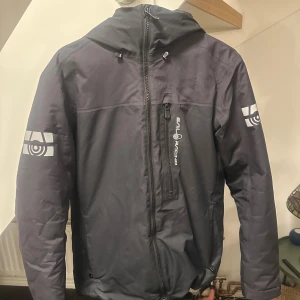 Svart jacka från Sail Racing - Snygg svart jacka från sailracing med justerbar nederkant med snörning för perfekt passform. Perfekt för blåsiga och kalla vinterdagar, den är helt perfekt utom ena sidan med snöringen som är innuti jackan det påverkas inte jackan iförsig eller utseendet.