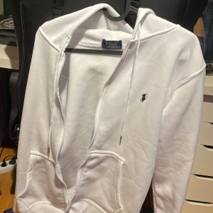 Vit hoodie från Polo Ralph Lauren - Vit hoodie från Polo Ralph Lauren med klassisk broderad logga på bröstet. Tröjan har huva med snörning, dragkedja framtill och två stora fickor. Mjuk och skön bomullskänsla, perfekt för chill dagar. Långärmad och stilren design.