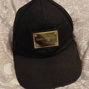 Svart keps från Dolce & Gabbana - Snygg svart keps från Dolce & Gabbana med en ikonisk metallplatta framtill där loggan står i guld. Kepsen är tillverkad i 100% bomull och har klassisk böjd skärm. Perfekt accessoar för att lyfta din streetwear-look. Nypris 2300.