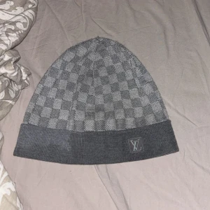 Grå Louis Vuitton Damier mössa - Snygg grå mössa från Louis Vuitton med klassiskt Damier-rutmönster. Mössan är stickad och har en vikt kant med LV-logga framtill. Perfekt accessoar för att lyfta din vinterstil med exklusiv känsla. Pris kan diskuteras billigare via snabbaffäre