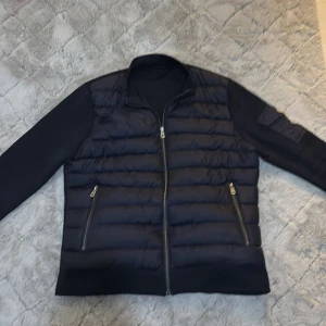   Moncler cardigan svart utan luva  - (Passar både M och S) Snygg svart pufferjacka från Moncler med quiltad front, släta ärmar och dragkedja. Jackan har två fickor med dragkedja framtill och en ikonisk Moncler-logga på ärmen. Materialmix av polyester och syntet, perfekt för höst och vinter.