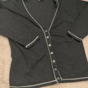 Mörkgrå kofta från Bon'a Parte - Mörkgrå kofta från Bon'a Parte med kontrasterande ljusa sömmar och knappar hela vägen framtill. Tillverkad i en mjuk mix av polyester och bomull. Perfekt för lager-på-lager och har långa ärmar med snygga detaljer vid muddar och kanter.