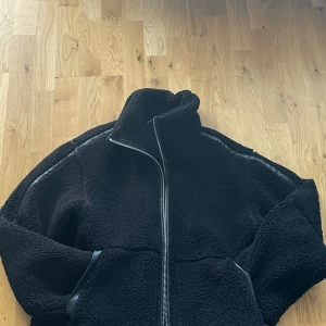 Svart teddyjacka med dragkedja - Svart teddyjacka med hög krage och hel dragkedja framtill. Jackan är i storlek xs men är oversized. Den sitter oversized på mig som är s/m och 176 cm lång. Jackan har detaljer i svart syntet längs ärmar och fickor, vilket ger en schysst kontrast mot det fluffiga materialet. Perfekt för kyliga dagar när du vill vara både varm och stilren.