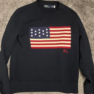 Mörkblå stickad tröja Polo Ralph Lauren - Säljer en mörkblå stickad tröja från Polo Ralph Lauren med amerikansk flagga framtill och RL-broderi. Tröjan har rund halsringning, ribbade muddar och lång ärm. Perfekt för dig som vill sticka ut med en klassisk och stilren look.