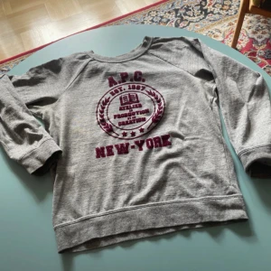 Grå sweatshirt från A.P.C. med tryck - Grå sweatshirt från A.P.C. med vinrött collegiatetryck på bröstet där det står 'A.P.C. New York'. Tröjan har rund halsringning, långa ärmar och är tillverkad i mjuk bomull. Perfekt för en chill och stilren look.