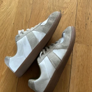 Vita och beige sneakers med mockadetaljer - Säljer ett par snygga sneakers från Maison Margiela i vitt och beige med bruna gummisulor. Skorna har paneler i mocka och skinn, klassisk rund tå och snörning. Perfekt för dig som gillar stilrena och trendiga sneakers med retrovibe.
