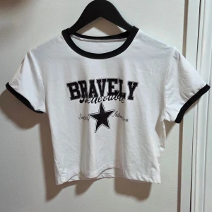 Vit croppad t-shirt med svart tryck, M - Snygg vit croppad t-shirt med svarta detaljer på krage och ärmslut. Framsidan har texten 'BRAVELY' och en stor svart stjärna med orden 'Simplicity Adventure'. T-shirten har en klassisk passform och är tillverkad i mjuk bomull.