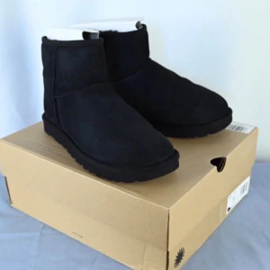Svarta UGG boots i mocka - Säljer ett par klassiska svarta UGG boots med rund tå och platt sula. Skorna är tillverkade i mjuk mocka och har en fluffig insida som håller fötterna varma. Perfekt för dig som gillar en chill och cozy stil under höst och vinter.