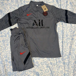 PSG grå långärmad träningströja Nike - Paris Saint-Germain långärmad träningströja i grått med svarta detaljer och röd Nike-logga. Tröjan har halv dragkedja, PSG-emblem på bröstet och diskret zebramönster på ärmarna. Tillverkad i lätt och funktionellt material, perfekt för fotbollsträning.