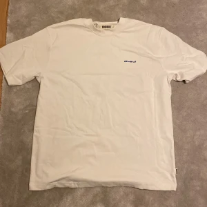 Vit Woodbird t-shirt med tigertryck - Vit t-shirt från Woodbird i 100% ekologisk bomull. Framsidan har en liten broderad logga i blått och baksidan har ett stort tryck med en blå tiger och grön text. Är öppen för ett lägre pris vid snabb affär. Om ni har några frågor skriv gärna!