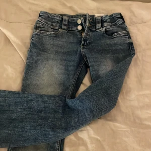 Blå bootcut jeans från H&M, stl 146 - Snygga blå bootcut jeans från H&M med låg midja och justerbar midja. Jeansen har klassisk femficksdesign, slitna detaljer och är tillverkade i bomull, polyester och elastan för en bekväm passform.