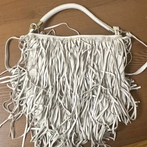 Vit axelväska med fransar i skinn - Snygg vit axelväska med långa fransar som ger en cool och bohemisk vibe. Väskan är tillverkad i mjukt skinn och har ett handtag samt axelrem med metalldetaljer. Perfekt för dig som vill sticka ut med en unik accessoar.