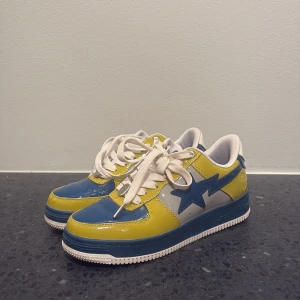 BAPE STA sneakers blå och gul - Säljer ett par BAPE STA sneakers i blankt skinn med cool färgkombo av mörkblått, gult och vitt. Skorna har klassisk snörning, perforerad tå och den ikoniska stjärnan på sidan. Perfekt för dig som vill sticka ut med streetwear-vibe. Använd en ända gång så praktiskt taget nya. Pris kan såklart diskuteras