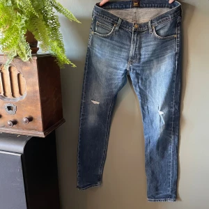 Blå Lee Luke jeans W32 L30 - Säljer ett par blå Lee Luke jeans med snygg slitning och små hål på knäna. Klassisk femficksmodell med gylf och knapp, samt Lee-logga på bakfickan. Jeansen har en smal passform och är tillverkade i bomullsdenim.