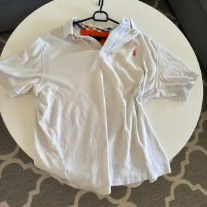 Vit piké från Polo Ralph Lauren XXL - Vit pikétröja från Polo Ralph Lauren i bomull, med klassisk krage och kort ärm. Detaljerad med broderad orange logga på bröstet och rutigt tyg på insidan av kragen. Perfekt för en clean och stilren look.
