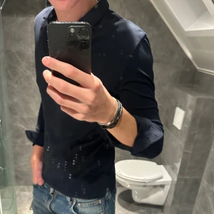 Mörkblå långärmad piké från J.Lindeberg - Säljer en stilren mörkblå långärmad piké från J.Lindeberg. Skjortan har lång ärm, klassisk krage och knappar framtill. Den är i riktigt bra skick. Jag är 188cm och den är för liten på mig. Hör av er vid funderingar;)