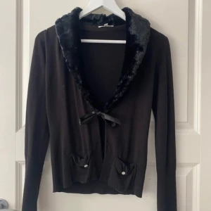 Svart kofta med fuskpäls - Supersöt svart kofta från Kookai med mjuk fuskpäls runt kragen och en söt rosett framtill. Två små fickor med dekorativa knappar ger extra detalj. Perfekt för att lyfta din outfit med en mysig och chic touch.