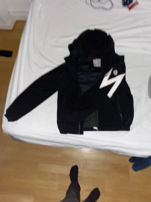 Svart vindjacka Moncler med huva - Svart vindjacka från Moncler med stor vit M-logga på vänstra ärmen. Jackan har huva, dragkedja framtill och två sidofickor med dragkedja. Materialet är lätt och syntetiskt, perfekt för blåsiga dagar. Snygg och modern design med Moncler-märke på ärmen.