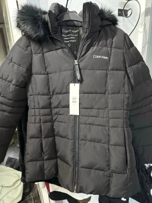 Svart pufferjacka från Calvin Klein - Svart pufferjacka från Calvin Klein med quiltad design och huva med snygg fuskpäls. Jackan har dragkedja framtill, två sidofickor och lättviktsisolering med Dupont Sorona. Perfekt för kalla dagar och har en stilren look med logga på bröstet.