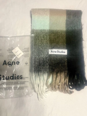 Rutig ullväst från Acne Studios - Säljer en rutig väst från Acne Studios i mjuk ullmix med stora blockrutor i beige, grått, svart, vitt och ljusgrönt. Västen har fransar nedtill och en tydlig Acne Studios-label framtill. Perfekt för dig som gillar stilrena detaljer och skandinavisk design.