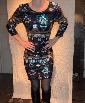 NY Svart paljettklänning nyårsklänning festklänning  med mönster CS - Snygg svart kortklänning med glittriga paljetter i geometriskt mönster i guld, lila och turkos. Klänningen har trekvartsärm och figurnära passform. Perfekt för dig som vill sticka ut med en unik look och älskar att glittra. Annat tyg i så inte sticksig av paljetterna! Jättefin
