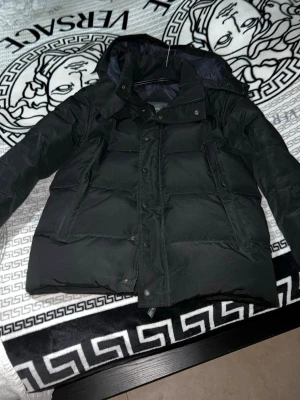 Svart pufferjacka från Canada Goose - Svart pufferjacka från Canada Goose med huva och snyggt ton-i-ton märke på ärmen. Jackan har dragkedja och knappar framtill, två sidofickor och quiltad design. Perfekt för kalla dagar och har en clean, modern look som funkar till streetwear. De med följer även päls och väska kommer samt att sätta på original lapparna.