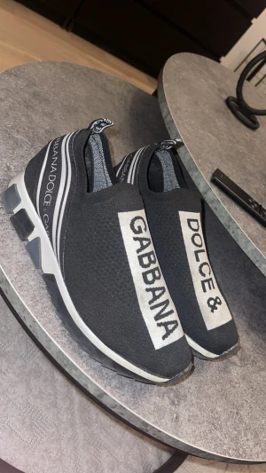 Dolce & Gabbana svarta sneakers - Svarta slip-on sneakers från Dolce & Gabbana med vit logotyptext på ovansidan och bakre delen. Snygg, sportig design med rund tå och platt sula. Tillverkade i stickad syntet med elastiska detaljer för skön passform. Perfekt för dig som gillar statement-plagg. Storlek 42 men de är stretchiga så passar även 43