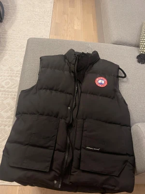 Svart dunväst från Canada Goose - Svart dunväst från Canada Goose med klassisk logga på bröstet. Västen har två stora fickor framtill, hög krage och dragkedja. Perfekt för kalla dagar och har en stilren, quiltad design. Snygg och funktionell för dig som gillar streetwear och outdoor. Storlek L, passar även M.
