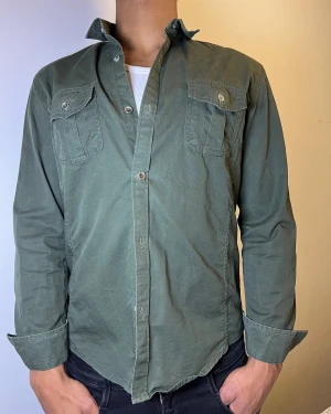 Grön skjorta med fickor och knappar - Snygg grön skjorta med klassisk krage och två bröstfickor med lock och knapp. Skjortan har långa ärmar som kan vikas upp och stängs med knappar framtill. Materialet är mjukt och känns som bomull, perfekt för en chill och stilren look.