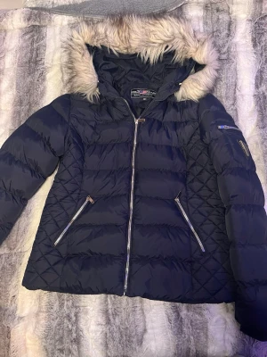 Mörkblå pufferjacka Polo Club - Snygg mörkblå pufferjacka från Polo Club Saint Vincent med fluffig fuskpäls på huvan. Jackan har quiltade detaljer på sidorna, två dragkedjeförsedda fickor och en cool patch på ärmen. Perfekt för kalla dagar med sin varma och stilrena look.