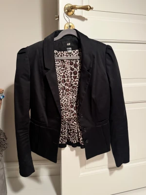 Svart kavaj från H&M, stl 38 - Svart kavaj från H&M i klassisk modell med snyggt fall och markerade axlar. Fodret har leopardmönster som ger en cool kontrast. Kavajen har knappar framtill och fickdetaljer. 🐆
