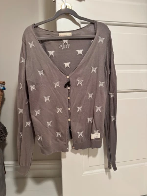 Grå kofta med broderade fjärilar - Supersöt grå kofta från Odd Molly med vita broderade fjärilar över hela tröjan. V-ringad med knappar framtill, snörning vid halsen och dragkedjor vid ärmsluten. Perfekt för dig som gillar unika detaljer och mjuka material❤️
