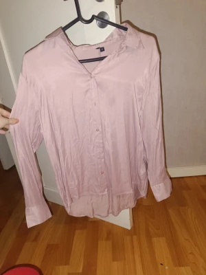 Ljusrosa skjorta med knappar - Säljer en ljusrosa skjorta från H&M med klassisk krage och knäppning framtill. Skjortan har långa ärmar med breda manschetter och är tillverkad i ett mjukt, tunt material som ger en skön känsla. Perfekt för dig som gillar stilrena och enkla plagg.
