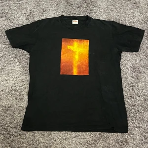 Supreme svart t-shirt med tryck - Supreme t-shirt i svart med ett stort, fyrkantigt tryck i orange och gult på bröstet. Trycket har ett abstrakt motiv som sticker ut. På ryggen finns Supreme-loggan och texten 'Andres Serrano' i gult. Klassisk passform och rund hals.