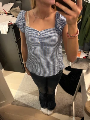 Blårutig blus med puffärmar - Supersöt blus i blåvitt rutigt mönster med puffiga ärmar som kan bäras både på axeln och offshoulder. Smockad rygg för skön passform och tre knappar framtill. Perfekt till jeans för en fräsch look. Säljer för det är inte riktigt min stil längre 