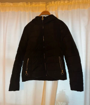 Svart pufferjacka med huva - Svart pufferjacka med huva och två fickor med snygga silverfärgade dragkedjor. Jackan har quiltad design och långa ärmar, perfekt för kyliga dagar. Stängs med dragkedja framtill och har en modern, enkel look.