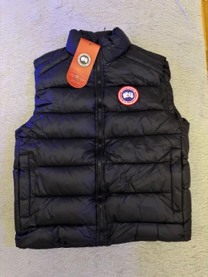 Svart dunväst från Canada Goose - Svart dunväst från Canada Goose med klassisk rund logotyp på bröstet. Västen har quiltad design, hög krage och dragkedja framtill. Materialet är polyester och dun, perfekt för kalla dagar. Snygg och stilren look med en normal passform.