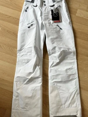 Vita täckbyxor från Stellar - Snygga vita täckbyxor från Stellar med flera dragkedjefickor och förstärkta partier vid knän och benslut. Byxorna har hög midja och justerbara hängslen för extra komfort. Perfekta för vinterns äventyr i backen.