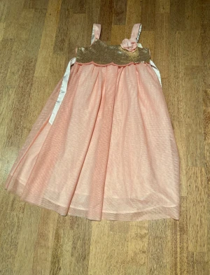 Rosa glittrig klänning från H&M - Supersöt rosa klänning från H&M med glittrigt guldigt liv, axelband och en dekorativ blomma framtill. Kjolen är lätt och luftig med ett tunt tyll-lager och vita band i midjan. Perfekt för dig som vill sticka ut lite extra!