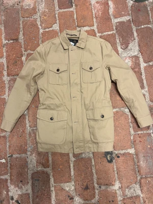 Beige overshirt från Morris, storlek S - Snygg beige overshirt från Morris med klassisk krage, fyra stora fickor med knappstängning och dragkedja framtill. Jackan har justerbar snörning i midjan och är tillverkad i slitstark bomull. Perfekt för dig som gillar stilrena och funktionella plagg. Orginal pris 2300 och säljs inte längre på webbplatsen 