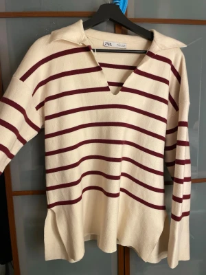 Randig polotröja från Zara i vinrött - Snygg polotröja från Zara i storlek S med bred krage och v-ringad hals. Tröjan är cremevit med vinröda horisontella ränder och har lång ärm. Materialet är mjukt och stickat, perfekt för dig som gillar en chill och trendig look.