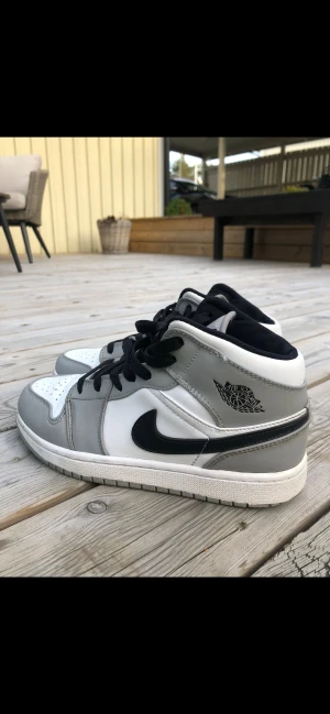 Nike Air Jordan 1 Mid grå/vit - Säljer ett par Nike Air Jordan 1 Mid i grått, vitt och svart. Klassisk siluett med svart swoosh, svarta skosnören och Air Jordan-logga på sidan. Ovandel i skinn och syntet, perforerad tå och vadderad plös för extra komfort. Riktigt snygga sneakers för dig som gillar streetstyle.